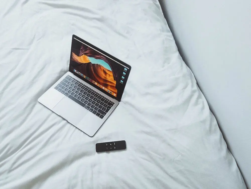 A laptop on a white bed with a remote.